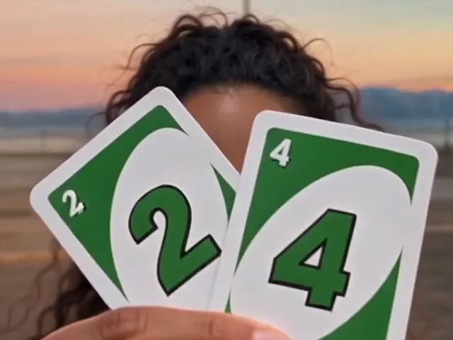 Close-up of a hand holding two green number cards to the camera, a large 2 in front and a large 4 behind it, with a woman's curly hair and a sunset sky behind them